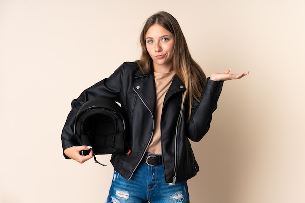 Joven mujer lituana sosteniendo un casco de motocicleta aislado en la pared beige con dudas mientras levanta las manos