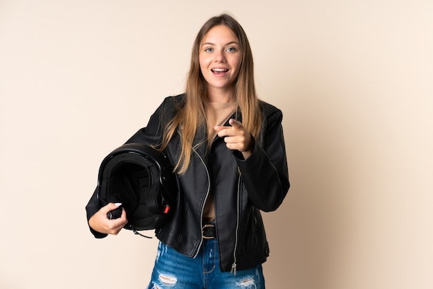 Joven mujer lituana sosteniendo un casco de motocicleta aislado en la pared beige sorprendido y apuntando hacia el frente