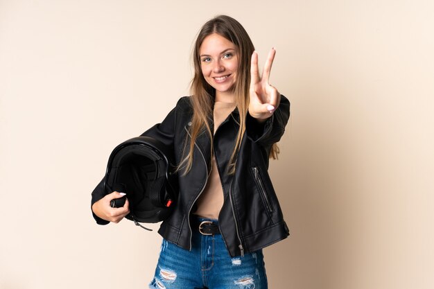 Joven mujer lituana sosteniendo un casco de motocicleta aislado sobre fondo beige sonriendo y mostrando el signo de la victoria