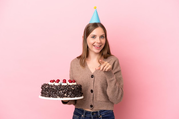 Joven mujer lituana sosteniendo pastel de cumpleaños aislado sobre fondo rosa sorprendido y apuntando hacia el frente