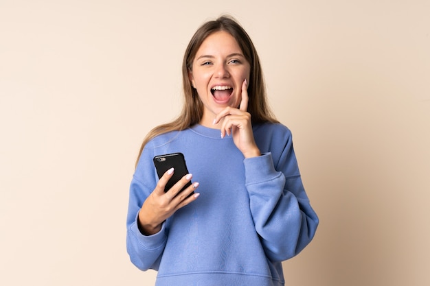 Foto joven mujer lituana a través de teléfono móvil aislado sobre fondo beige gritando con la boca abierta