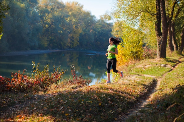 Joven mujer llena de vida haciendo ejercicio, haciendo su rutina corriendo en el hermoso parque de la ciudad a principios de otoño, un paisaje increíble con río y bosque al fondo