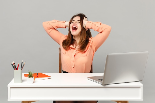 Joven mujer loca gritando manteniendo los ojos cerrados, poniendo las manos en la cabeza sentada, trabajando en el escritorio con una computadora portátil aislada en un fondo gris. Concepto de estilo de vida de carrera empresarial de logro. Simulacros de espacio de copia.