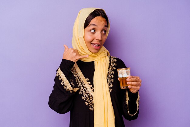 Joven mujer marroquí sosteniendo un vaso de té aislado sobre fondo púrpura mostrando un gesto de llamada de teléfono móvil con los dedos.