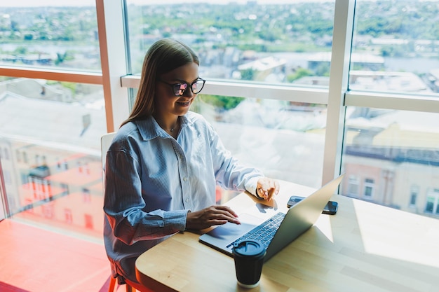 Joven mujer milenaria con anteojos de protección ocular clásicos que se conectan a Internet WIFI en una computadora portátil en un espacio de coworking para buscar información web en netbooks digitales mujer europea chateando