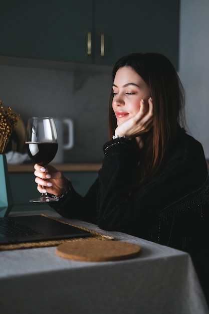 Joven mujer milenaria con videollamadas en una computadora portátil y bebiendo vino, usa la tecnología para comunicarse con amigos o familiares