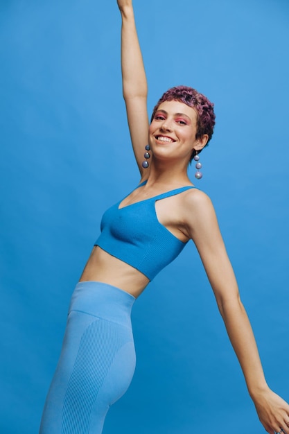 Joven mujer de moda atlética con cabello teñido y corte de pelo corto posando y bailando en ropa deportiva azul sonriendo y mirando a la cámara sobre un fondo monocromático azul