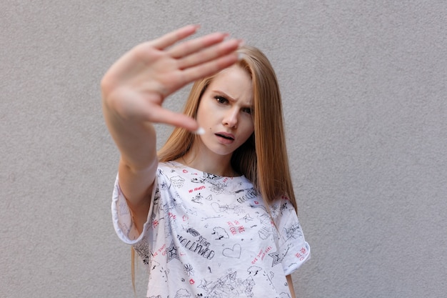 Joven mujer de moda europea con expresión de enojo insatisfecho sosteniendo la mano en gesto de parada tratando de cubrir su rostro de la cámara, aislado sobre fondo blanco. Es difícil ser modelo famoso