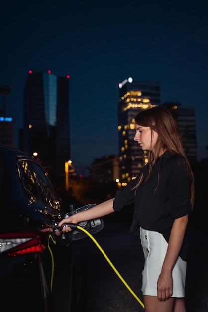 Joven mujer moderna cargando un coche eléctrico ev en la ciudad por la noche