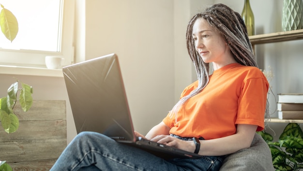 Joven mujer moderna con rastas está cómodamente sentada en una bolsa de silla en casa y usando una computadora portátil