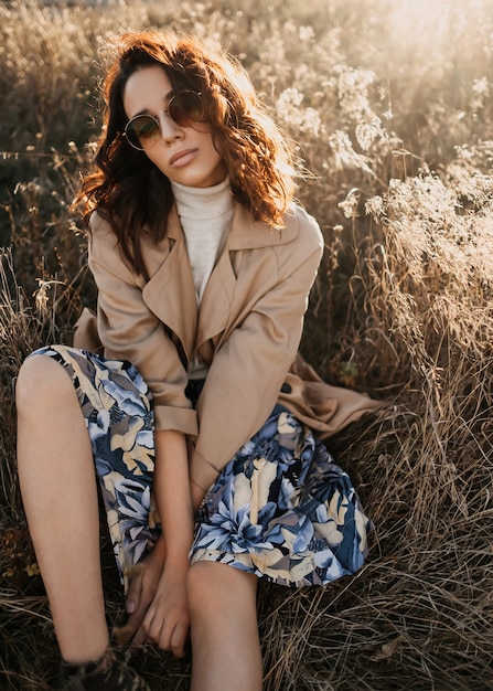 Joven mujer moderna sentada en un campo, con gafas de sol al atardecer