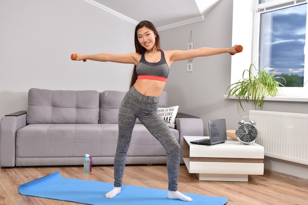 Joven mujer morena asiática en ropa deportiva haciendo ejercicios en casa con dos pesas naranjas.