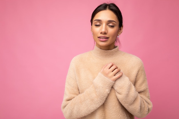 Joven mujer morena bonita linda positiva con emociones sinceras vistiendo suéter beige aislado sobre fondo rosa con espacio vacío y sosteniendo la mano en el pecho con los ojos cerrados.