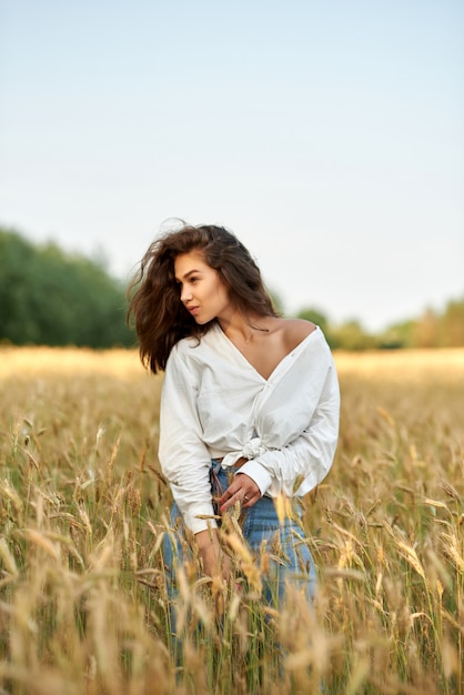 Joven mujer morena con camisa blanca y pantalones vaqueros azules sobre un fondo de campo de trigo dorado