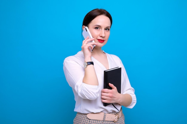 Una joven mujer morena con un diario está hablando por teléfono sobre un fondo azul.