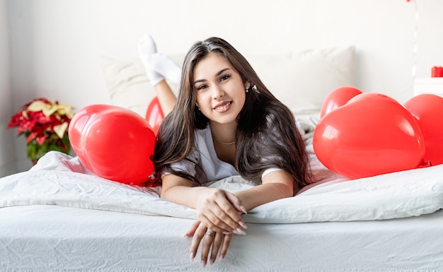 Joven mujer morena feliz acostado en la cama con globos en forma de corazón rojo