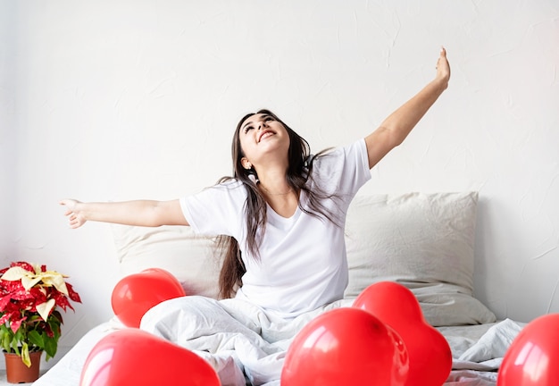 Joven mujer morena feliz sentada despierta en la cama con globos en forma de corazón rojo