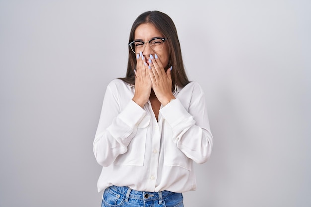 Joven mujer morena con gafas riendo y avergonzada riendo cubriendo la boca con las manos chismes y concepto de escándalo