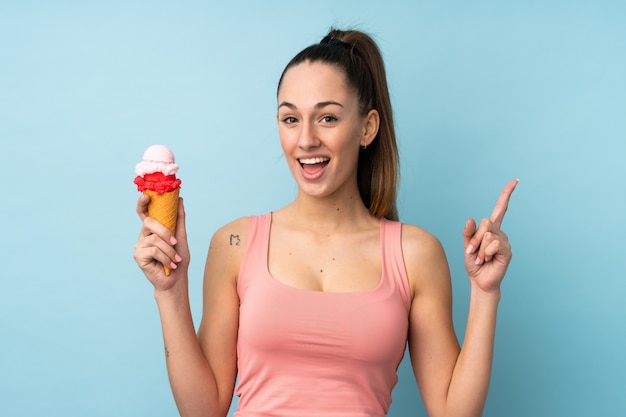 Joven mujer morena con un helado de cucurucho sobre pared azul aislado apuntando hacia arriba una gran idea