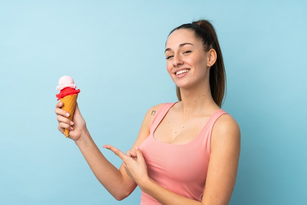 Joven mujer morena con un helado de cucurucho sobre pared azul aislado y apuntando