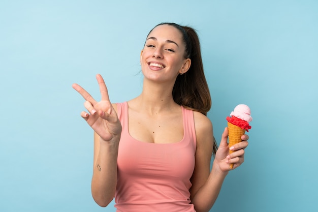 Joven mujer morena con un helado de cucurucho sobre pared azul aislado sonriendo y mostrando el signo de la victoria