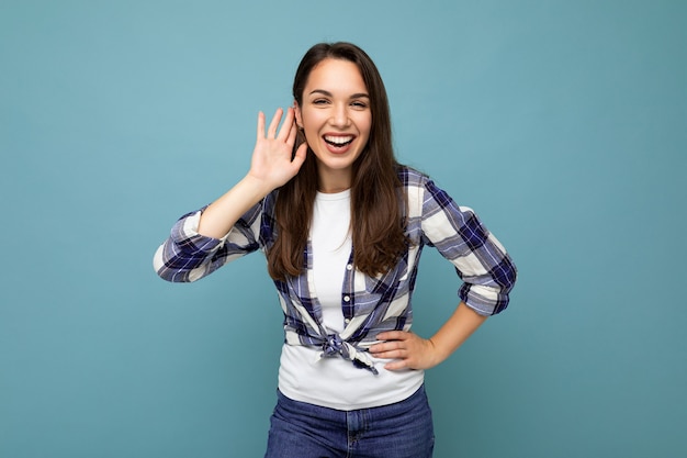 Joven mujer morena hermosa positiva con emociones sinceras con camisa a cuadros de moda de pie aislado sobre fondo azul con espacio vacío y escuchando algo poniendo la mano en la oreja