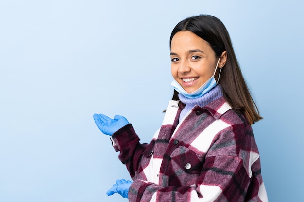 Joven mujer morena de raza mixta que protege del coronavirus con una máscara y guantes sobre una pared azul aislada extendiendo las manos hacia un lado para invitar a venir