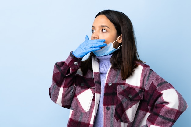 Joven mujer morena de raza mixta que protege del coronavirus con una máscara y guantes sobre la pared azul aislada haciendo un gesto de sorpresa mientras mira hacia un lado