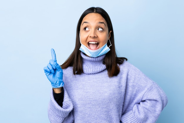 Joven mujer morena de raza mixta que protege del coronavirus con una máscara y guantes sobre una pared azul aislada con la intención de darse cuenta de la solución mientras levanta un dedo