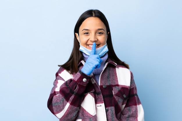 Joven mujer morena de raza mixta que protege del coronavirus con una máscara y guantes sobre una pared azul aislada que muestra un gesto de silencio poniendo el dedo en la boca
