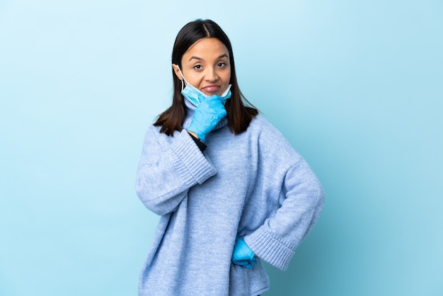 Joven mujer morena de raza mixta que protege del coronavirus con una máscara y guantes sobre pensamiento de pared azul aislado