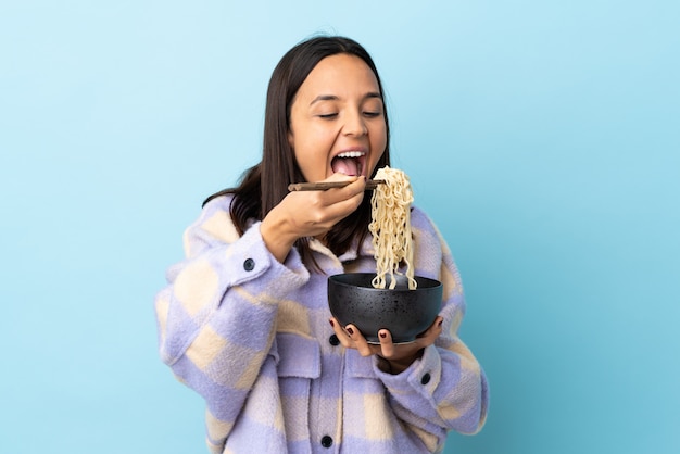 Joven mujer morena de raza mixta sobre pared azul aislado sosteniendo un tazón de fideos con palillos y comiéndolo