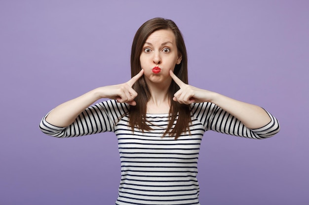 Joven mujer morena con ropa informal a rayas posando aislada en un retrato de estudio de fondo morado violeta. Concepto de estilo de vida de las personas. Simulacros de espacio de copia. Señalar con el dedo índice las mejillas hinchadas.