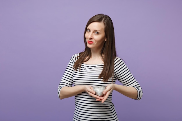 Joven mujer morena con ropa informal a rayas posando aislada en un retrato de estudio de fondo morado violeta. Gente emociones sinceras concepto de estilo de vida. Simulacros de espacio de copia. Sostén el corazón de plata plateada