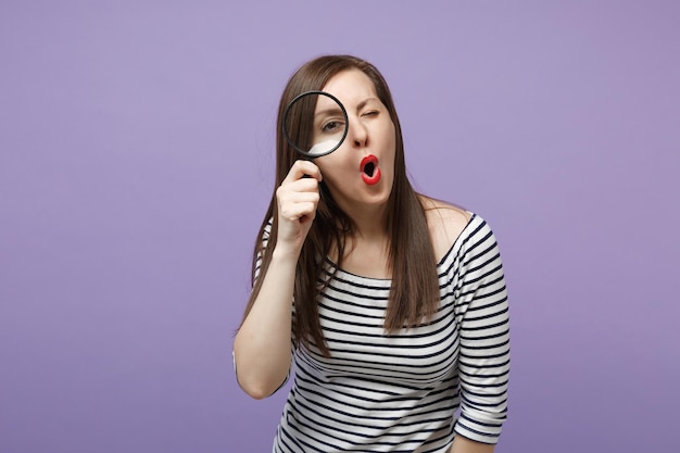 Joven mujer morena con ropa informal a rayas posando aislada en un retrato de estudio de fondo morado violeta. Gente emociones sinceras concepto de estilo de vida. Simulacros de espacio de copia. Sostenga la lupa.