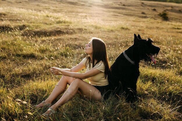 Joven mujer morena sentada en un campo al lado de un gran perro negro de raza gigante schnauzer