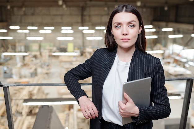 Joven mujer morena seria gerente de ventas de la fábrica de muebles contemporáneos de pie delante de la cámara y mirándote