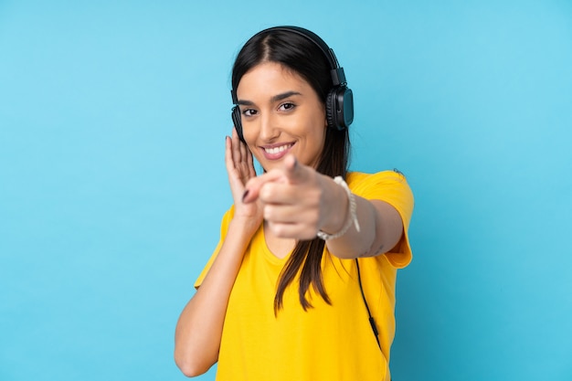 Joven mujer morena sobre pared azul aislada escuchando música y apuntando hacia el frente