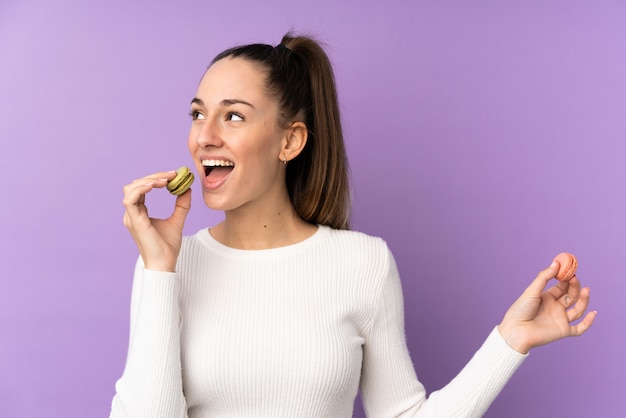 Foto joven mujer morena sobre pared púrpura aislado sosteniendo coloridos macarons franceses y comiéndolo