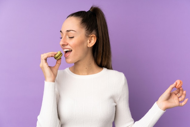 Joven mujer morena sobre pared púrpura aislado sosteniendo coloridos macarons franceses y comiéndolo