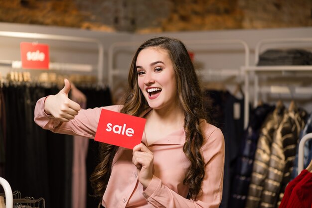 Joven mujer morena sonriente mostrando el pulgar hacia arriba y mantenga el cartel rojo con la palabra de venta