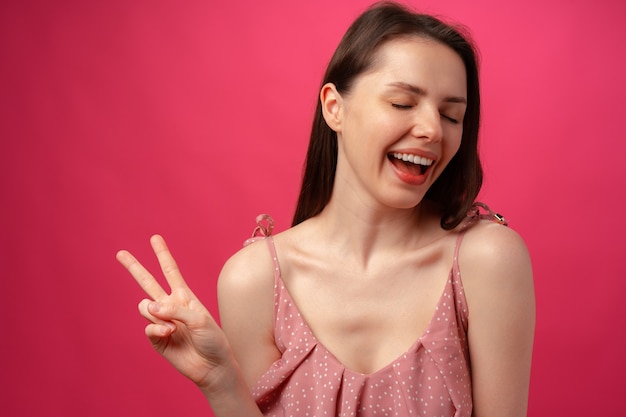 Joven mujer morena sonriente mostrando el signo de la victoria contra el fondo de color rosa