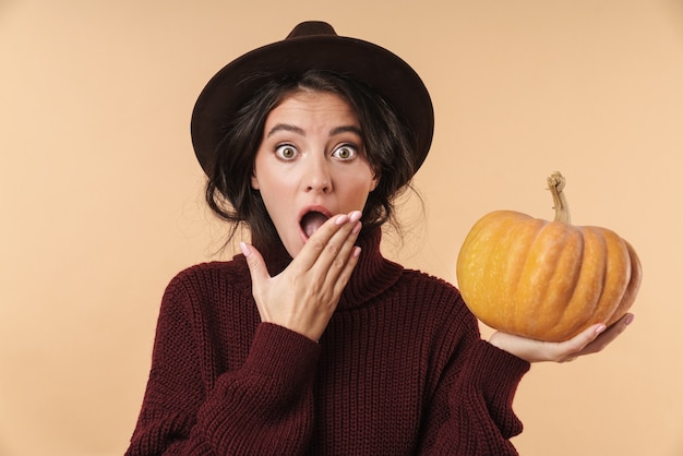 joven mujer morena sorprendida aislada sobre la pared de la pared de color beige con calabaza.