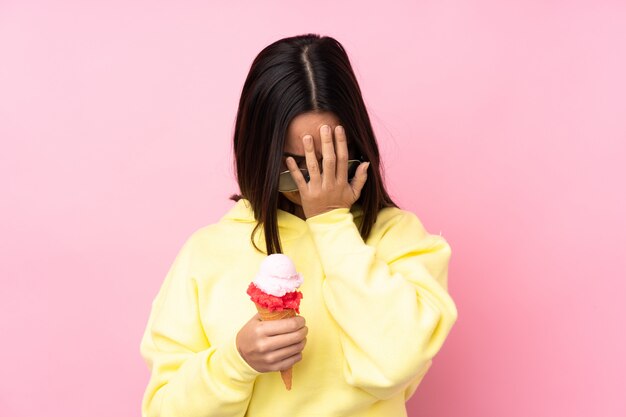 Joven mujer morena sosteniendo un helado de cucurucho sobre pared rosa aislado con expresión cansada y enferma