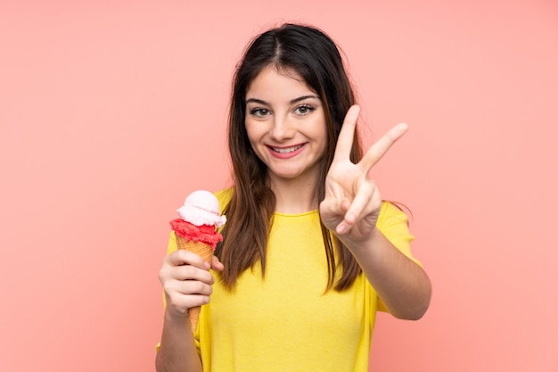 Joven mujer morena sosteniendo un helado de cucurucho sobre pared rosa aislado sonriendo y mostrando el signo de la victoria