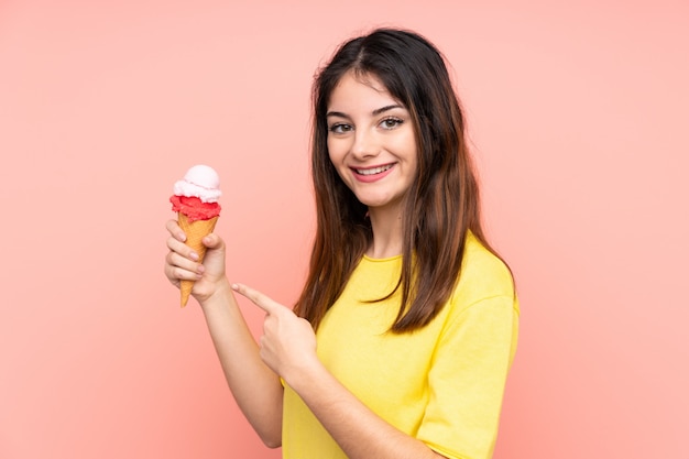 Joven mujer morena sosteniendo un helado de cucurucho sobre pared rosa y apuntando