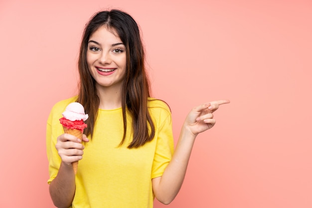 Joven mujer morena sosteniendo un helado de cucurucho sobre pared rosa sorprendido y apuntando con el dedo hacia un lado