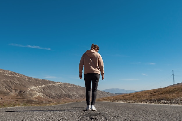 Joven mujer morena con sudadera con capucha de color se encuentra a lo largo de la carretera de asfalto vacía entre altas colinas bajo un cielo azul en un día soleado