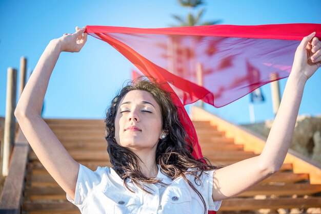 Joven mujer morena vestida de blanco contra el cielo azul bufanda roja cerrar ojos feliz placer en el viento