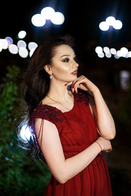 Joven mujer morena con un vestido rojo con un tren posando en un parque nocturno